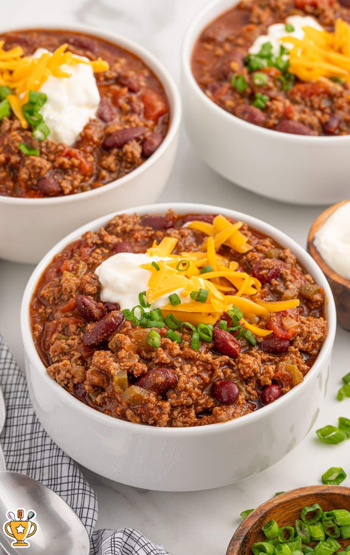Three white bowls of crockpot chili on a light tabletop, showing chunky ground beef, kidney beans, tomatoes, and green pepper, topped with shredded cheddar, a dollop of sour cream, and sliced green onions.