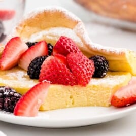 Slice of dutch baby pancake on a white plate, dusted with powdered sugar and topped with strawberries and blackberries, with a fork holding a bite in the foreground on a bright white tabletop.