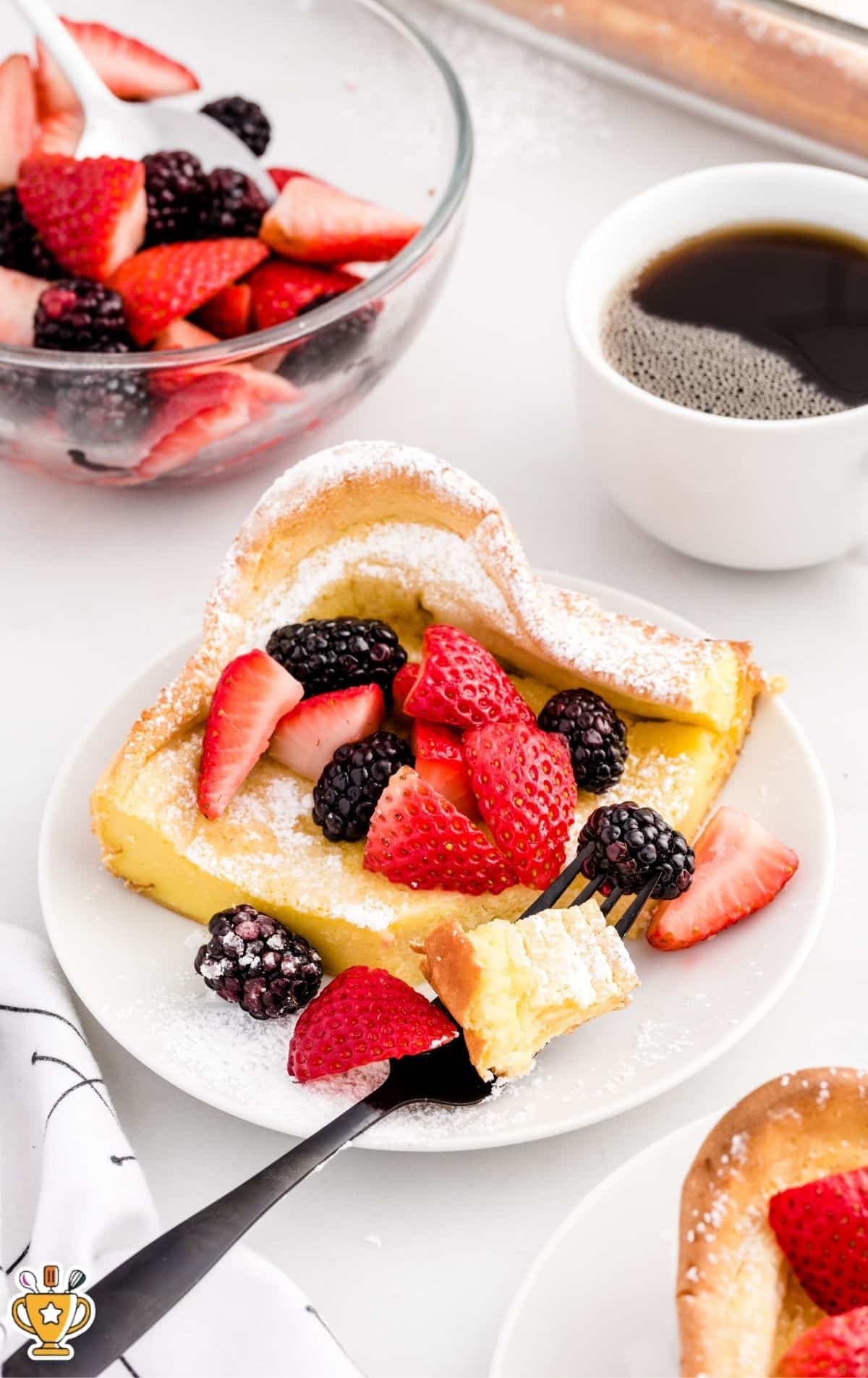 Plated slice of dutch baby pancake dusted with powdered sugar and topped with strawberries and blackberries, with a fork holding a bite; background includes a bowl of mixed berries and a cup of coffee on a bright white surface.