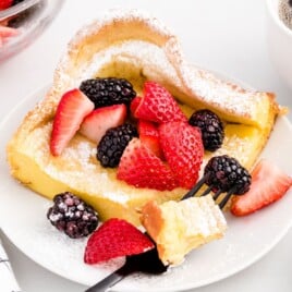 Close-up of a slice of dutch baby pancake on a white plate, heavily dusted with powdered sugar and topped with fresh strawberries and blackberries, with a fork lifting a bite.