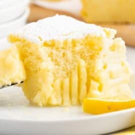 Fork lifting a bite of Lemon Custard Cake from a white plate, showing the creamy custard center and soft cake layer, with a lemon slice on the plate and more cake squares blurred in the background.