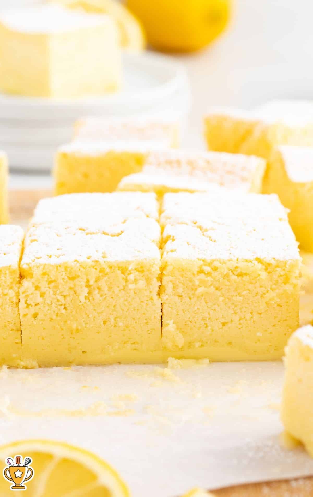 Close-up of several Lemon Custard Cake squares dusted with powdered sugar, showing the layered custard and cake texture, with additional squares softly blurred in the background.