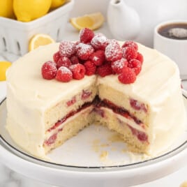Whole Lemon Raspberry Cake on a white cake stand with a large slice removed, showing raspberry filling and cream cheese frosting between lemon cake layers, topped with a mound of powdered-sugar-dusted raspberries with lemons and a coffee cup in the background.
