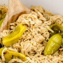 Close-up of Mississippi chicken in a slow cooker with shredded chicken and whole pepperoncini peppers, with a wooden spoon stirring in the background.