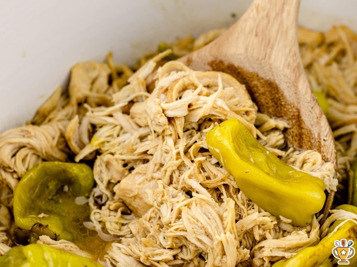 Slow cooker Mississippi chicken with shredded chicken, pepperoncini peppers, and au jus seasoning in a rich buttery sauce, shown with a wooden spoon lifting tender, juicy meat.