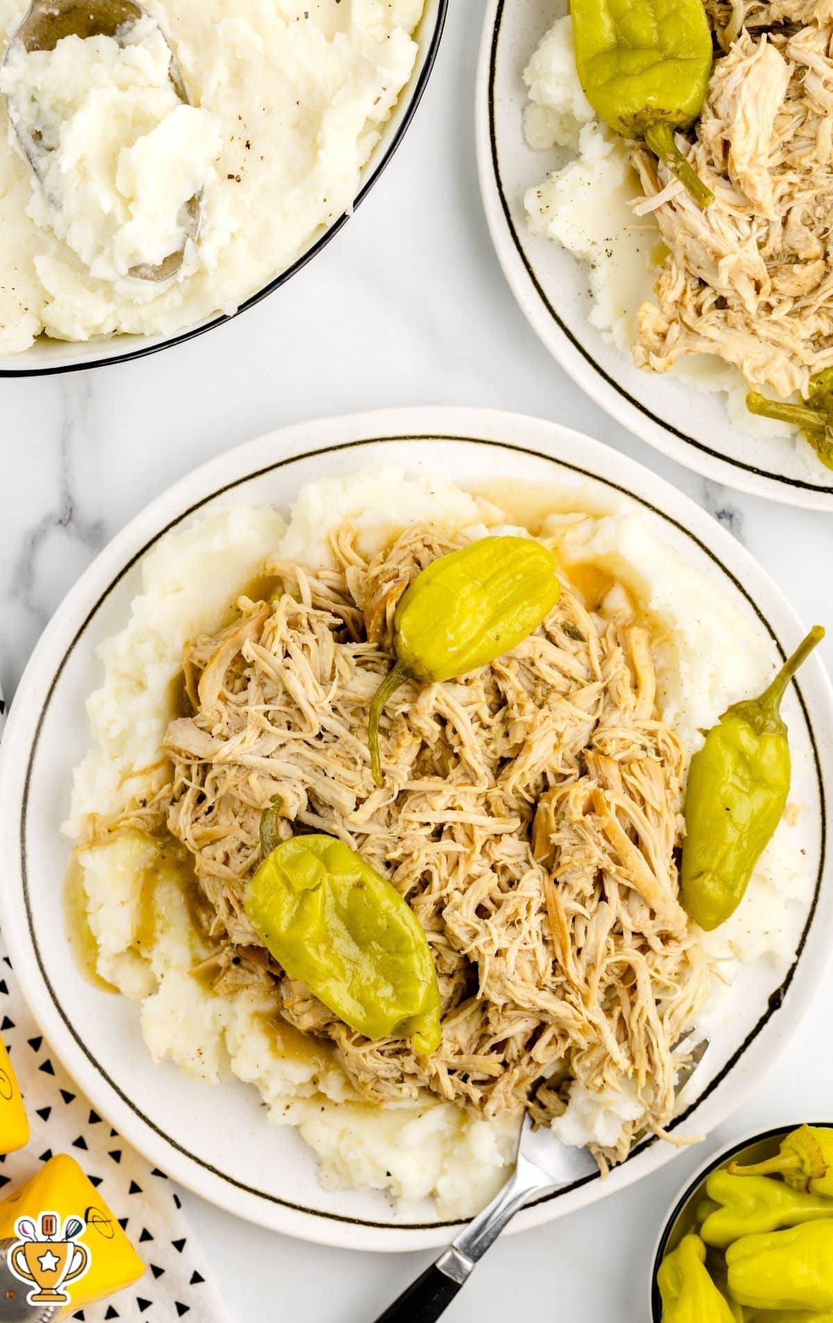 Overhead view of Mississippi chicken served over mashed potatoes on a white plate with pepperoncini peppers and gravy, with a spoon and extra plates on a light marble surface.