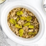 Overhead shot of Mississippi chicken in a white slow cooker, showing shredded chicken in au jus with whole pepperoncini peppers on top.