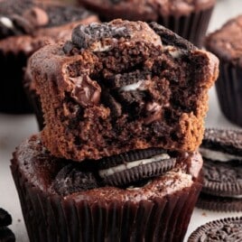 Close-up of rich chocolate Oreo muffins in dark brown liners, with one muffin split open and stacked on another to show Oreo cookie chunks and chocolate chips inside; whole Oreo cookies and more muffins are scattered in the background on a light surface.
