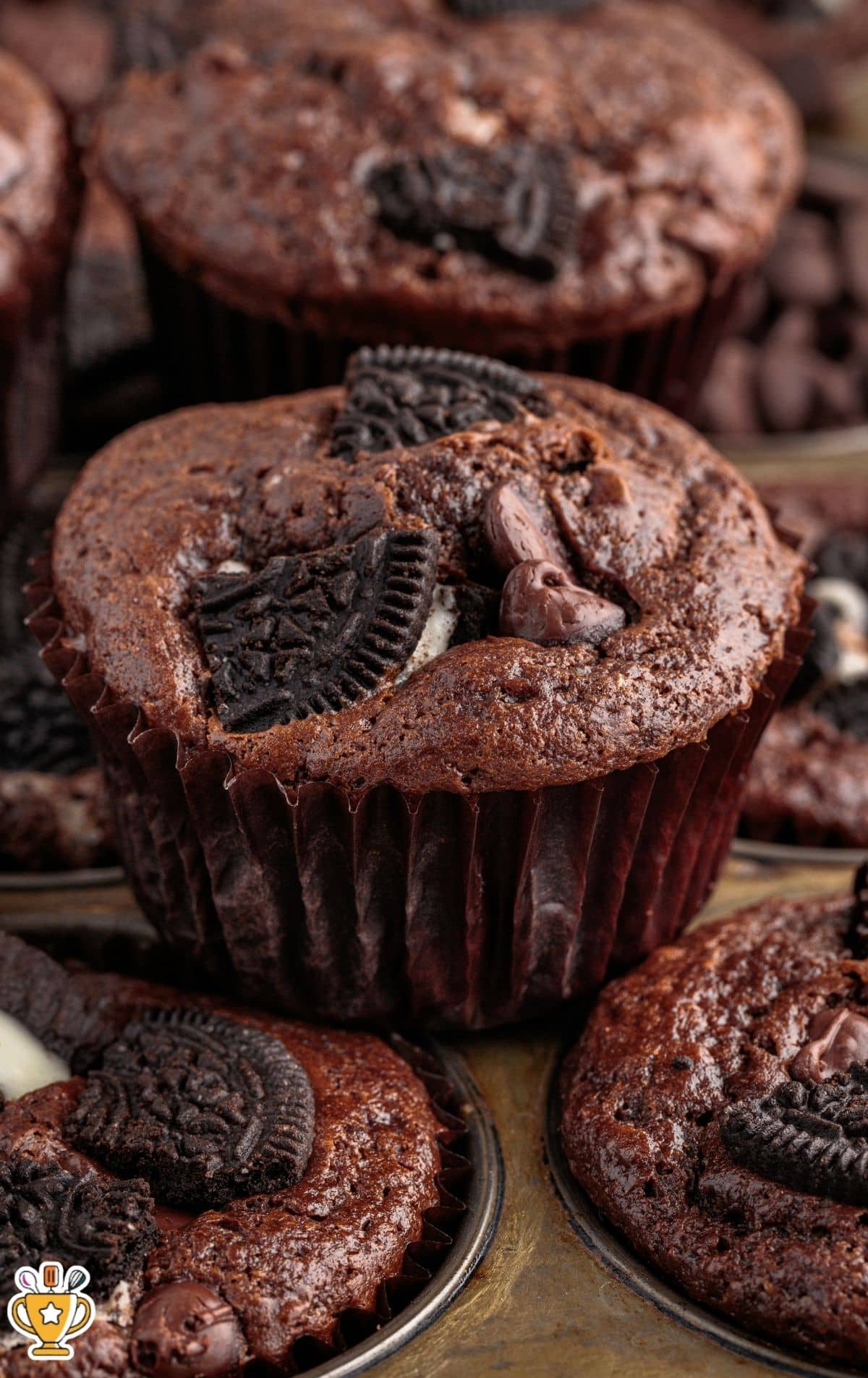 Tight close-up of several baked chocolate Oreo muffins in a muffin tin, each topped with Oreo cookie pieces and chocolate chips in dark brown liners, with cookie crumbs around the pan and a small trophy-and-utensils logo in the corner.