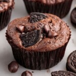 Close-up of a baked chocolate Oreo muffin in a dark brown liner, topped with Oreo cookie pieces and chocolate chips, with other muffins and Oreo cookies softly blurred in the background on a light surface.