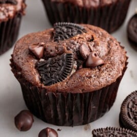 Close-up of a baked chocolate Oreo muffin in a dark brown liner, topped with Oreo cookie pieces and chocolate chips, with other muffins and Oreo cookies softly blurred in the background on a light surface.