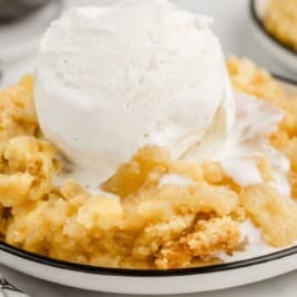 Bowl of Pineapple Dump Cake topped with vanilla ice cream, showing the moist pineapple base and crumbly yellow cake topping. A baking dish of the dessert sits in the background, highlighting the simple, classic pineapple dump cake texture.