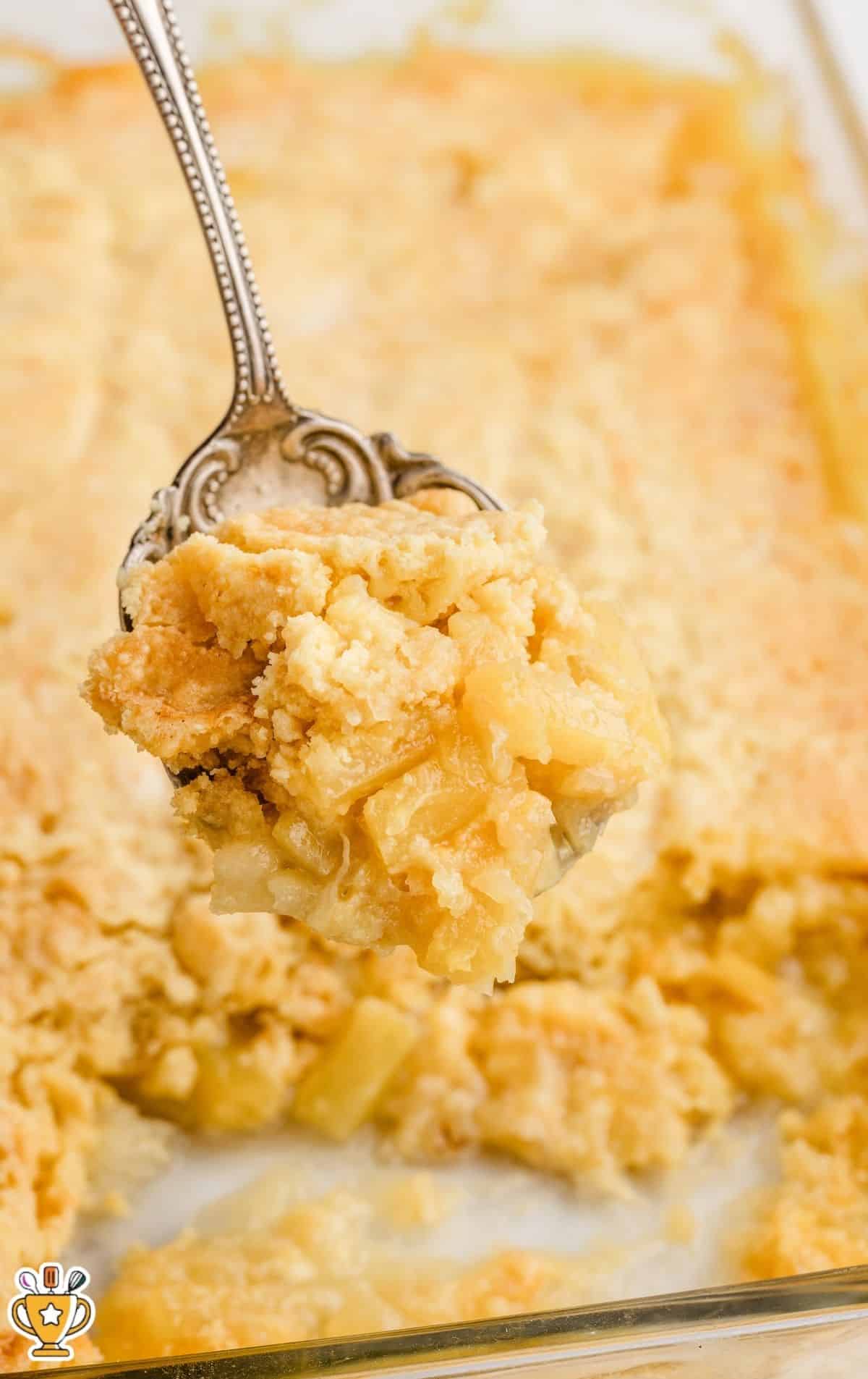 Close-up of a spoon lifting a warm, gooey serving of Pineapple Dump Cake from a baking dish, showing tender pineapple chunks and buttery golden cake crumble topping.
