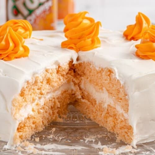 Whole orange soda cake on a clear glass cake stand, frosted in smooth white icing with orange frosting swirls on top, with a large slice missing to show the orange crumb and creamy white filling.