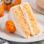 Close-up of a slice of orange soda cake with white frosting on a white plate, topped with an orange frosting rosette, with Crush Orange soda cans softly blurred in the background.