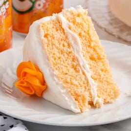 Close-up of a slice of orange soda cake with white frosting on a white plate, topped with an orange frosting rosette, with Crush Orange soda cans softly blurred in the background.