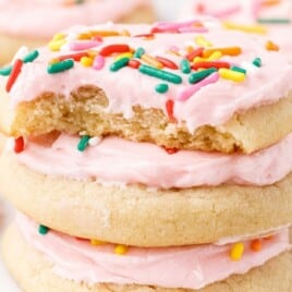 Stack of soft sugar cookies frosted with pale pink icing and topped with rainbow sprinkles, with the top cookie showing a bite, and more frosted sprinkle cookies blurred in the background.