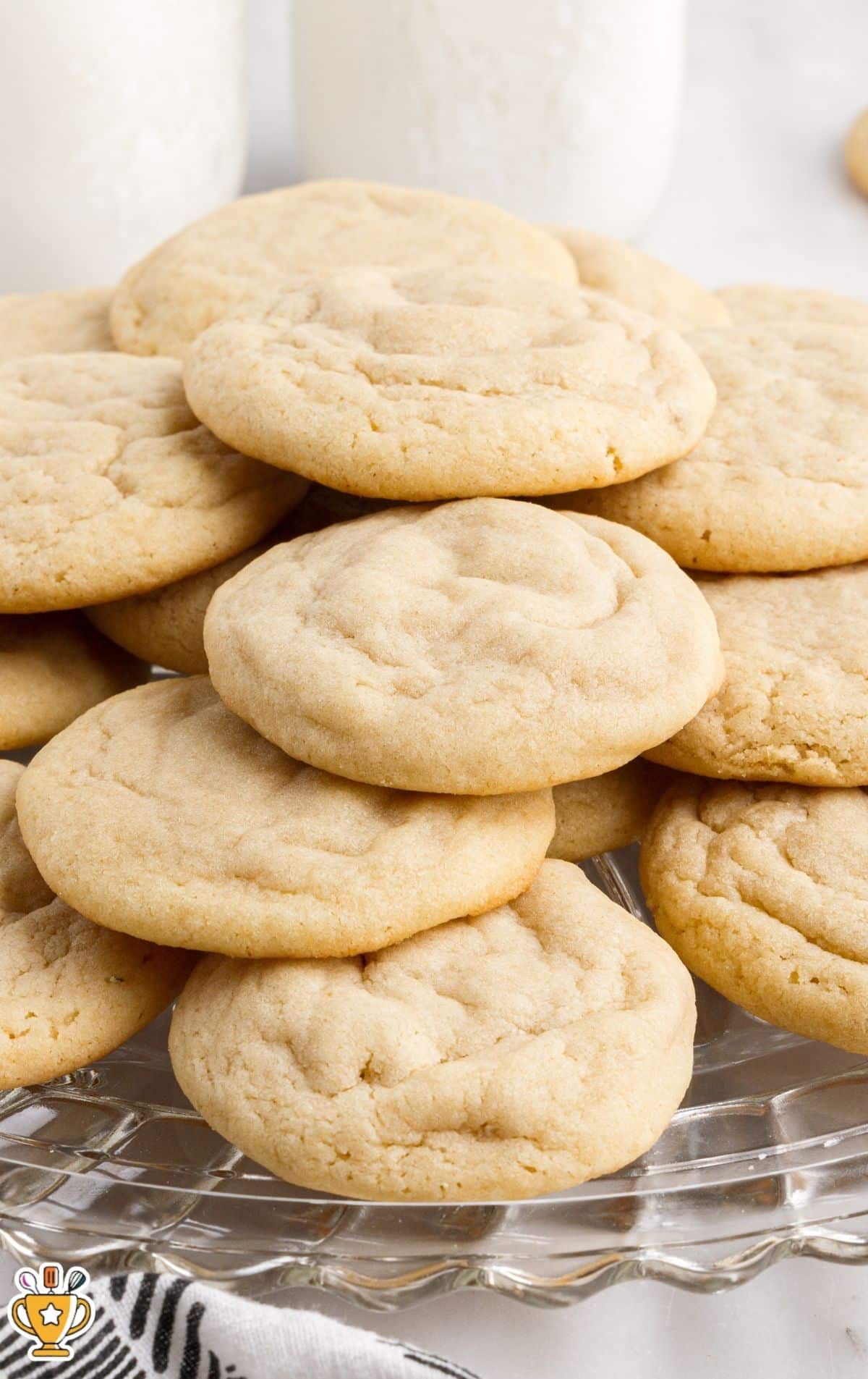 Close-up of plain soft sugar cookies stacked on a clear glass serving plate, with white jars and a neutral kitchen background, highlighting the lightly golden edges and tender cookie texture.