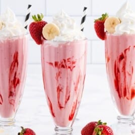 Three tall strawberry banana smoothies in vintage sundae glasses with strawberry swirls, topped with whipped cream, banana slices, strawberries, and black-and-white striped straws, styled on a white surface with bananas and fresh strawberries in the background.
