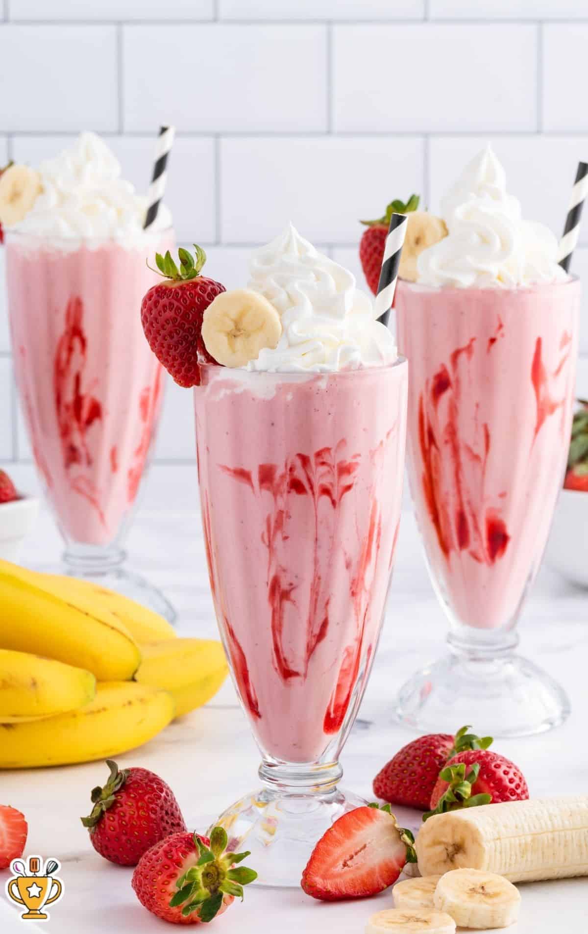 Vertical photo of three strawberry banana smoothies in tall sundae glasses with red strawberry swirls, topped with whipped cream, banana slices, strawberries, and striped straws, with bananas and strawberries arranged around the glasses on a bright white background.