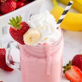 Strawberry banana smoothie in a glass mason jar mug, topped with whipped cream and garnished with a fresh strawberry and banana slice, with a black-and-white striped straw and a pink smoothie drip down the jar; strawberries and bananas are blurred in the background.
