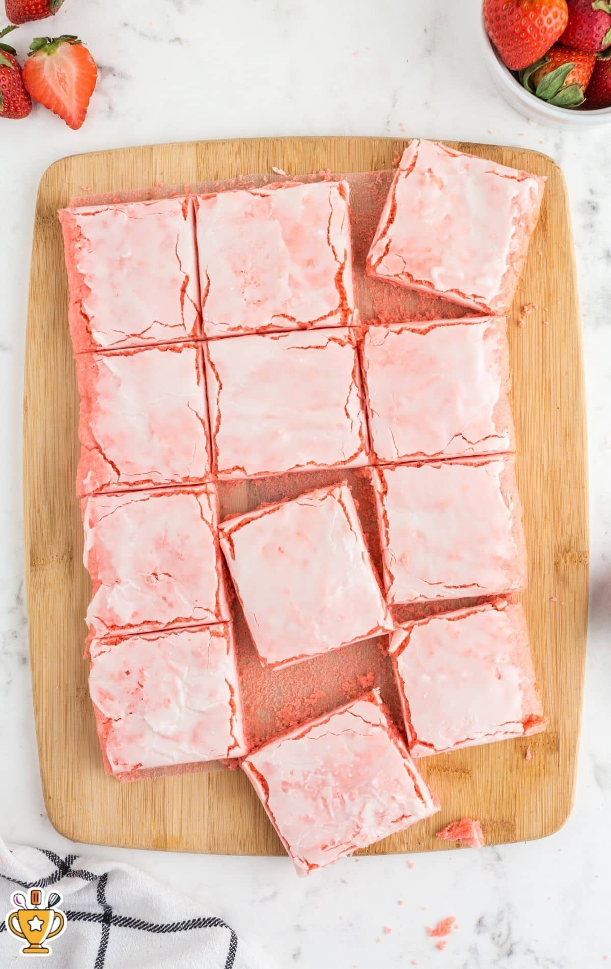 Overhead view of strawberry brownies cut into squares on a wooden cutting board over a white marble surface, with fresh strawberries in the corners.