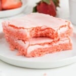Two strawberry brownies stacked on a white plate with a bite taken out of the top bar, showing a soft pink crumb and crackly pale pink icing.