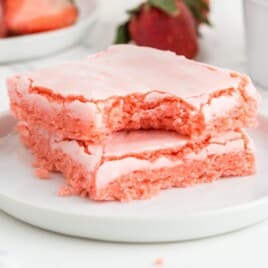 Two strawberry brownies stacked on a white plate with a bite taken out of the top bar, showing a soft pink crumb and crackly pale pink icing.