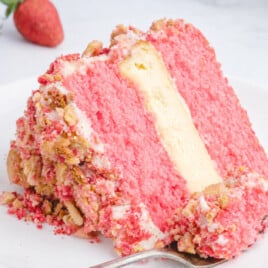 Horizontal close-up of a Strawberry Crunch Cake slice with bright pink strawberry cake layers, a thick creamy vanilla cheesecake center, whipped topping, and golden cookie crumb coating on a white plate with fresh strawberries and a fork on a marble countertop.