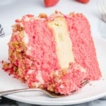 Close-up of a Strawberry Crunch Cake slice with bright pink strawberry cake layers, a thick vanilla cheesecake center, whipped cream frosting, and golden cookie crumb coating on a white plate with a fork.