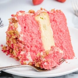 Close-up of a Strawberry Crunch Cake slice with bright pink strawberry cake layers, a thick vanilla cheesecake center, whipped cream frosting, and golden cookie crumb coating on a white plate with a fork.