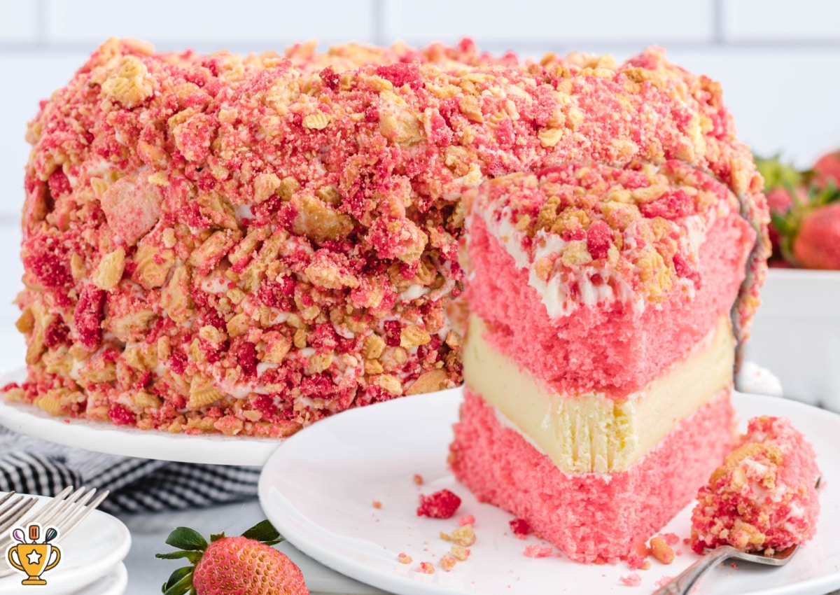 Strawberry crunch cake with pink strawberry cake layers, a creamy cheesecake center, and a strawberry cookie crumb coating, shown with a sliced piece on a white plate and fresh strawberries nearby.