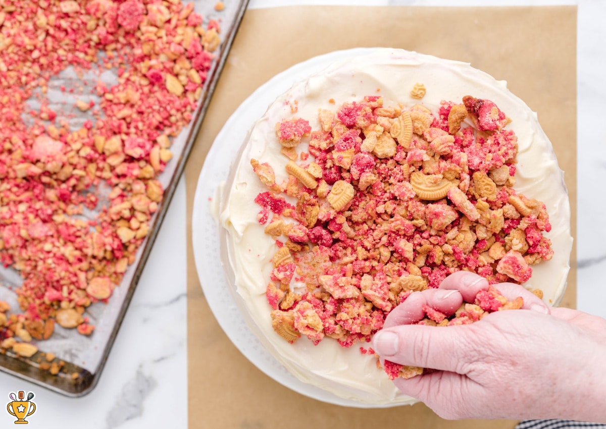 Overhead view of hands pressing strawberry crunch cookie crumbs onto a frosted strawberry crunch cake, with extra crumbs on a parchment-lined baking sheet nearby.