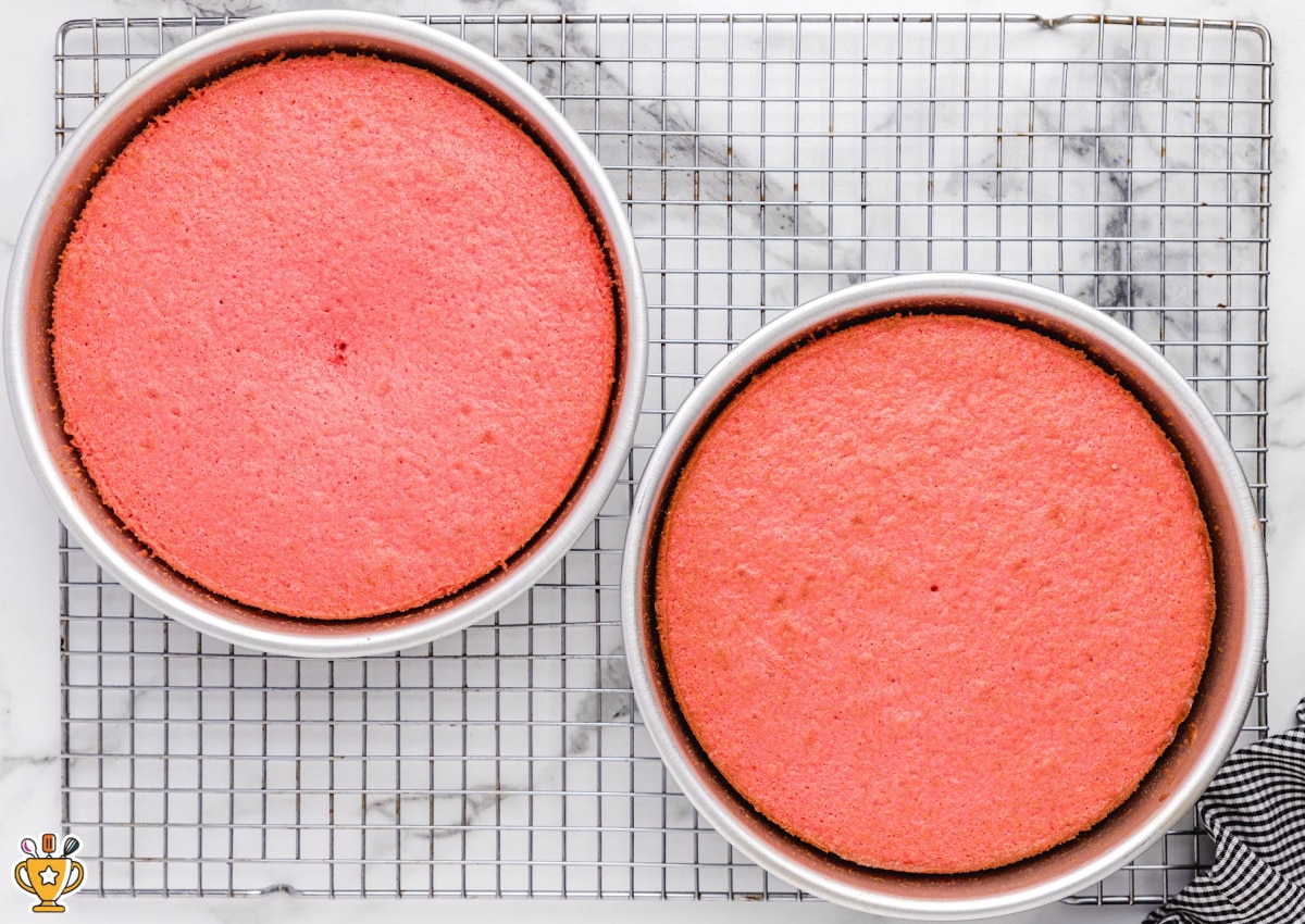 Two strawberry cakes baked and cooling in round cake pans. 
