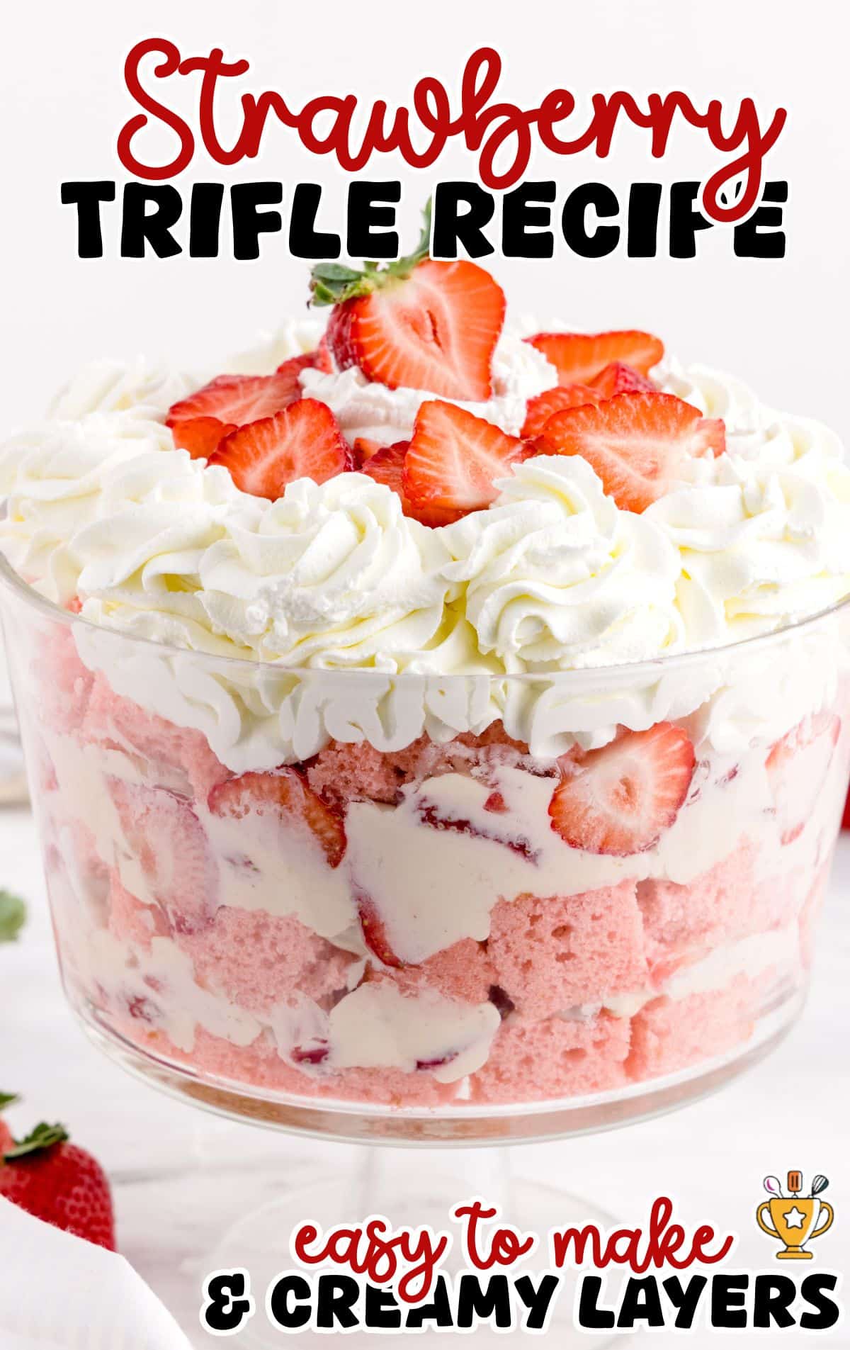 Strawberry trifle layered dessert in a clear glass bowl featuring pink strawberry cake, creamy cheesecake mixture, and fresh sliced strawberries, finished with piped whipped cream and strawberry garnish on top. Image text reads “Strawberry Trifle Recipe” and “easy to make & creamy layers” on a clean white background.