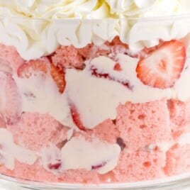 Strawberry Trifle layered in a clear glass trifle bowl with cubes of strawberry cake, sliced fresh strawberries, and creamy cheesecake filling, topped with piped whipped cream and halved strawberries.