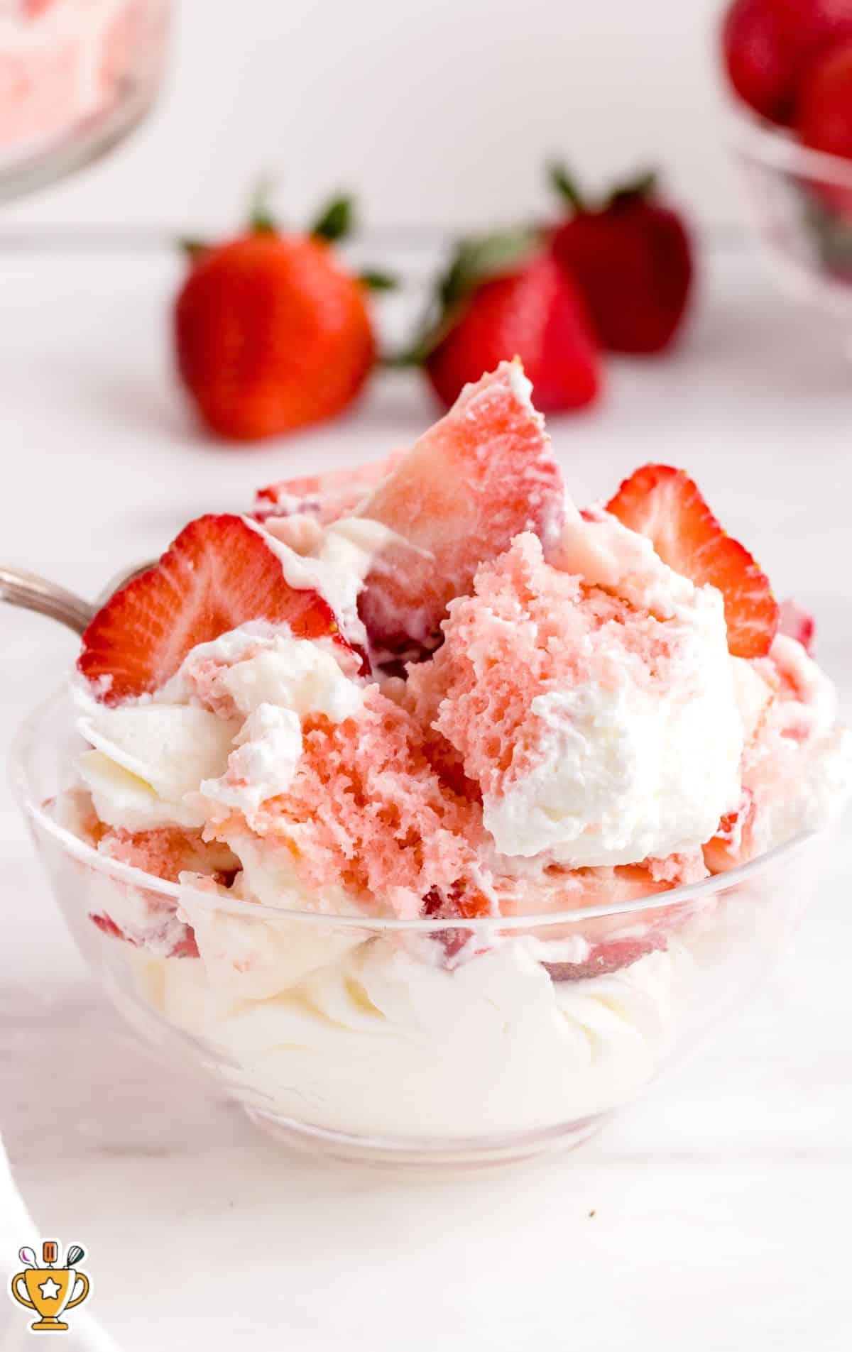 Close-up of a serving of Strawberry Trifle in a small glass bowl, showing fluffy strawberry cake pieces, fresh strawberry slices, and creamy whipped filling with a spoon lifting a bite.