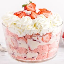 Recipe card image for Strawberry Trifle showing a large clear glass bowl with fluffy strawberry cake, sliced strawberries, and creamy filling, topped with piped whipped cream and fresh strawberry halves.