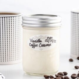 Mason jar of homemade vanilla coffee creamer labeled “Vanilla Coffee Creamer” centered on a white surface, with two patterned mugs in the background and coffee beans scattered to the side.