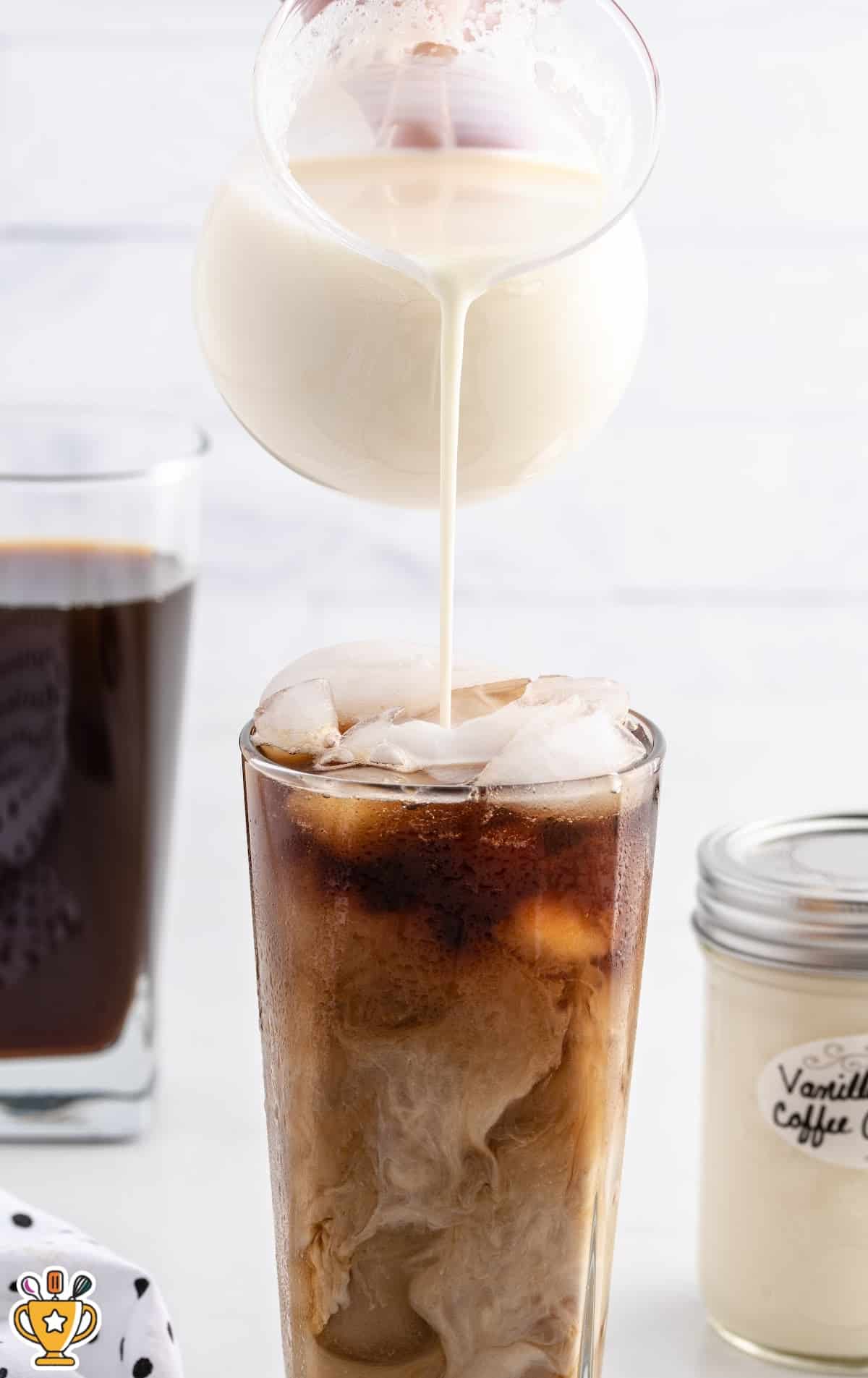 Creamy homemade coffee creamer pouring into a tall glass of iced coffee, with a small jar of vanilla coffee creamer partially visible on the side.