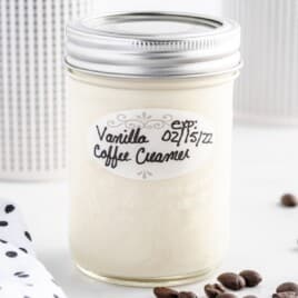 Mason jar filled with homemade vanilla coffee creamer, labeled “Vanilla Coffee Creamer” with an expiration date, styled on a bright white background with a few coffee beans.