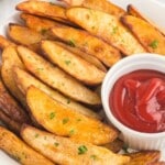 Crispy golden air fryer potato wedges served on a white plate with chopped parsley and a small bowl of ketchup for dipping.