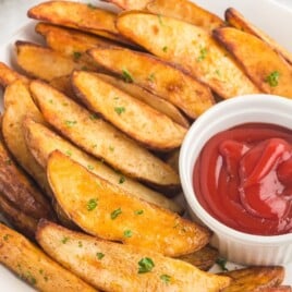 Crispy golden air fryer potato wedges served on a white plate with chopped parsley and a small bowl of ketchup for dipping.