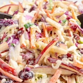 Close-up of creamy apple slaw tossed in a glass bowl with wooden salad servers, featuring shredded coleslaw mix, sliced red apples, carrots, green onions, and dried cranberries in a sweet creamy dressing.