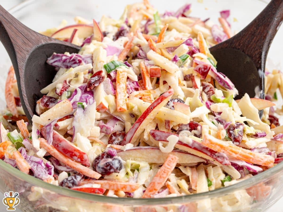 Close-up of Apple Slaw in a glass mixing bowl with wooden salad spoons, showing crunchy cabbage, red apple matchsticks, carrots, green onions, and dried cranberries coated in creamy dressing.