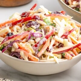 Bowl of creamy Apple Slaw with shredded cabbage, red apple slices, carrots, green onions, and dried cranberries in a sweet creamy dressing, served on a gray surface with extra slaw in the background.
