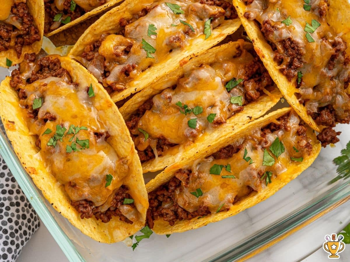 Baked tacos arranged upright in a glass baking dish, filled with seasoned ground beef and topped with melted cheddar-jack cheese, lightly garnished with chopped cilantro.