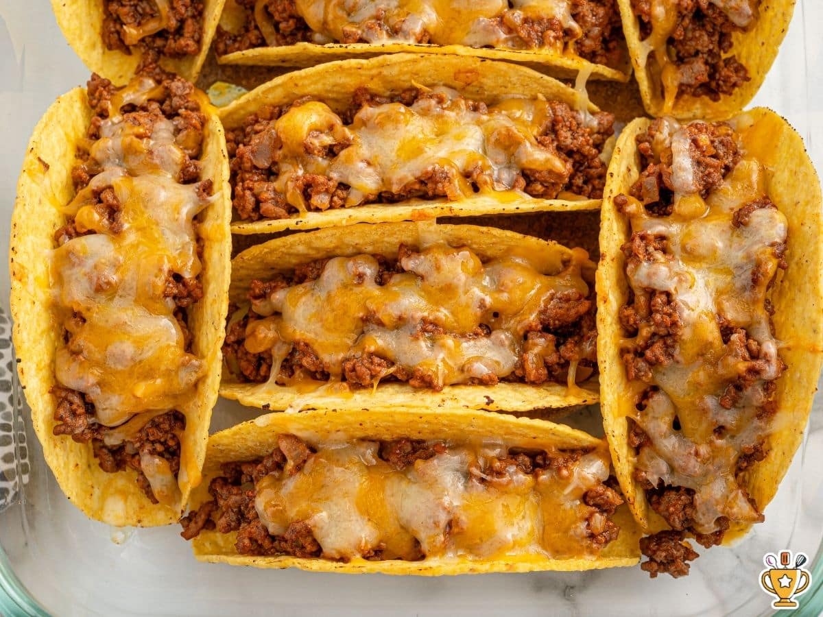 Baked hard shell tacos arranged upright in a glass baking dish, filled with seasoned ground beef and topped with melted cheddar-jack cheese, with crisp edges and evenly melted tops.