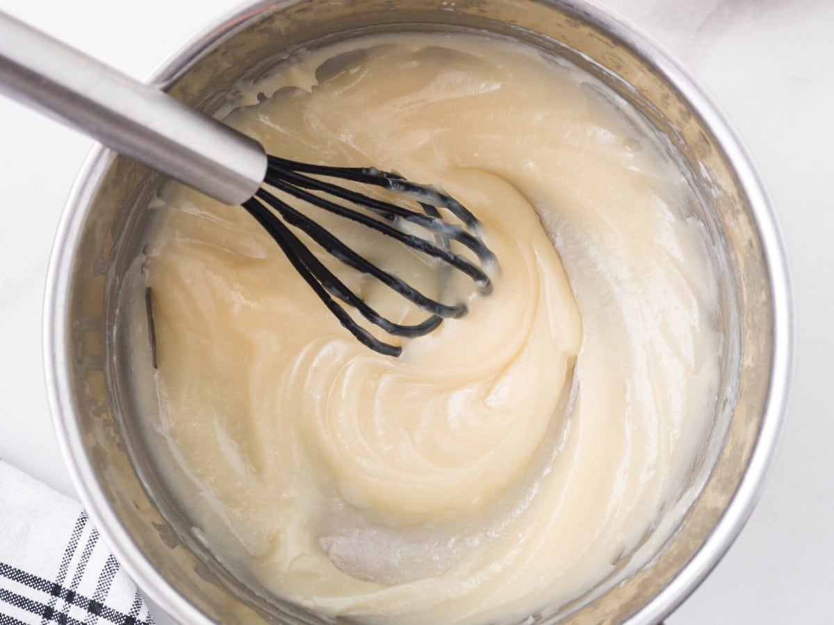 Close-up of a saucepan with a thick, pale ermine mixture being whisked, showing a smooth, pudding-like consistency forming on the stove.