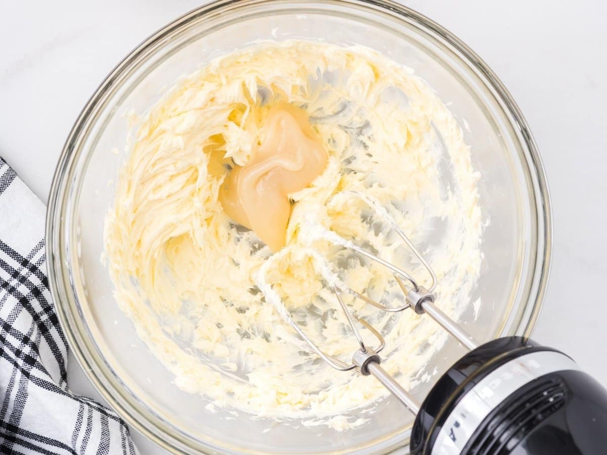 Creamed butter in a glass mixing bowl with sweetened condensed milk being added, partially blended with an electric hand mixer, showing a soft, fluffy texture in the early stage of ermine frosting preparation.